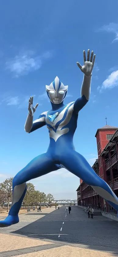 横浜赤レンガ倉庫前の広場にAR合成された青銀カラーのウルトラマン（アグル系）の巨大立像。青空と赤レンガ建物を背景にダイナミックなポーズで立つ全身像