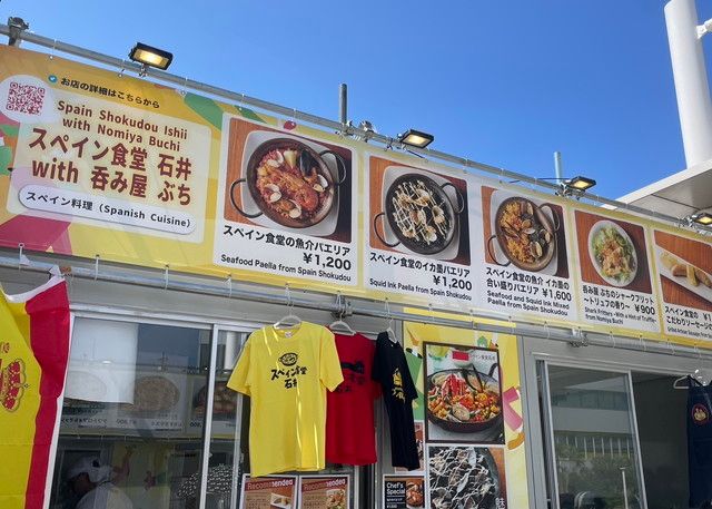スペイン食堂 石井 with 呑み屋 ぶちのブース外観。パエリアなどのメニュー写真が大きく掲げられた店頭
