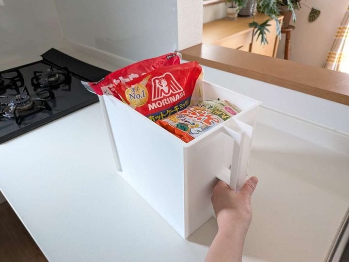 白いキッチンカウンターの上で、取っ手付きの白い収納ボックスにホットケーキミックスやうどんなどの食品ストックを立てて収納し、手で取っ手を引いている写真