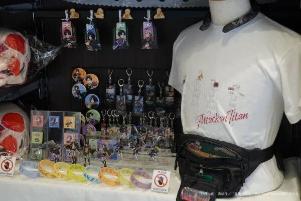 進撃の巨人コラボグッズ販売ブースの陳列写真。アクリルキーホルダー・缶バッジ・ラバーブレスレット・アクリルスタンドなど複数種のグッズと、Attack on TitanロゴプリントTシャツ＋ウエストバッグを着用したマネキンを展示