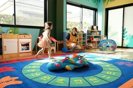 キッズルーム内でスタッフが子どもと遊ぶ託児施設の様子。カラフルなマットと玩具棚を備えた広い室内で女児とスタッフが映っている