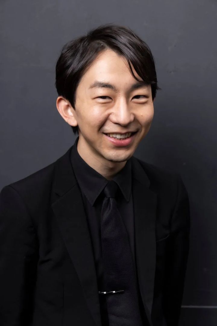 お笑い芸人・リップグリップ岩永圭吾さんの宣材写真風ポートレート。黒スーツ・黒シャツ・黒ネクタイ姿で笑顔を見せる男性の上半身が灰色背景に映る。