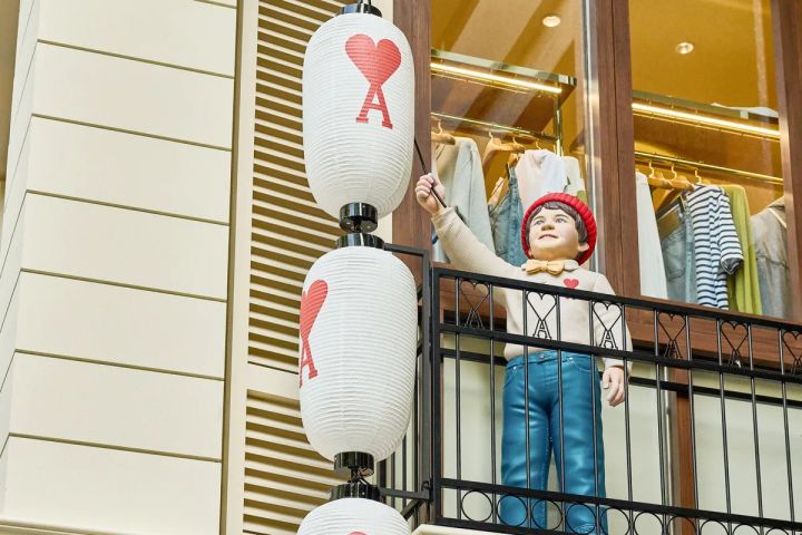Ami Parisのハートロゴ入り白い提灯3連と、赤いベレー帽・蝶ネクタイ・ジーンズ姿の男の子フィギュアのバルコニーディスプレイのクローズアップ。背後にジャケット・Tシャツ等の衣類ハンガーが並ぶ