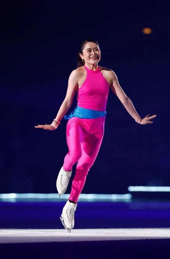 ピンクのノースリーブコスチュームにブルーのウエストリボンをつけた女性スケーターがリンク上でジャンプ直後の着氷姿勢をとるパフォーマンスシーン
