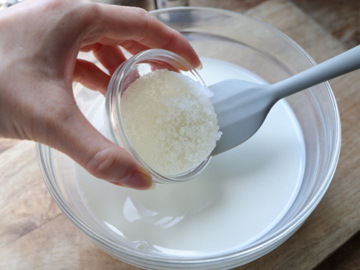 牛乳の入った大きなガラスのボウルに、ふやかしたゼラチンが入った小さなガラスの容器をシリコンヘラを添えて加えようとしている手元写真