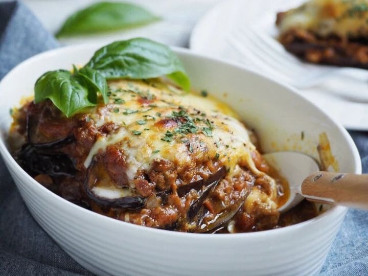 白い深皿にキーマカレーとなすが層に重なり、上面にチーズ・刻みパセリ・バジルが乗りスプーンを添えた写真
