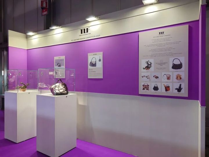 東京レザーフェア（TLF）のコンテスト展示ブース全景。紫の壁面にレザーデザインコンペティション2025の入賞作品パネルが貼られ、白台座の展示ケースに受賞革製品バッグ2点が陳列されている
