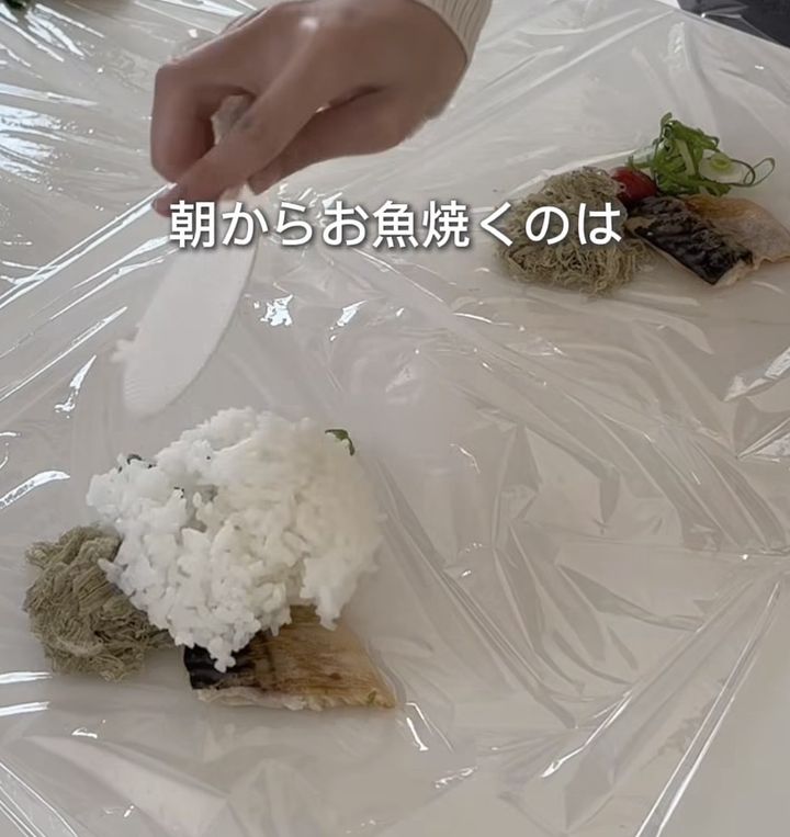 A hand holds a spatula over a sushi-style piece with white rice on top placed on plastic wrap, with greens and other toppings nearby; Japanese caption reads '朝からお魚焼くのは'.