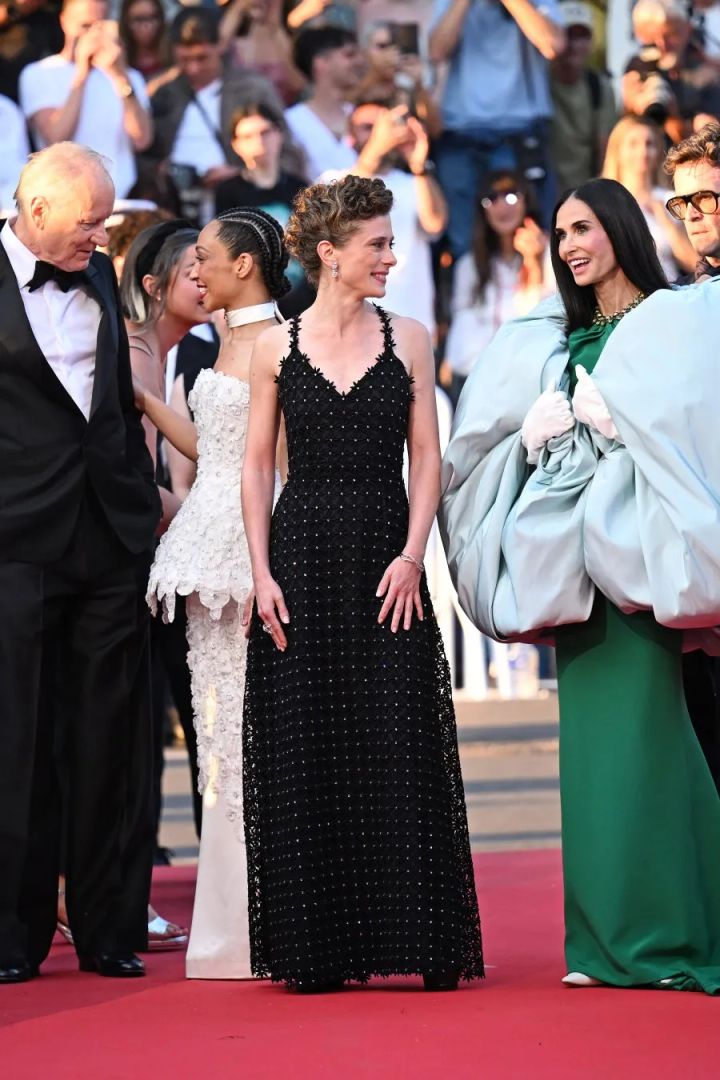 カンヌ映画祭レッドカーペットにてローラ・ワンデルが刺繍入りブラックのAラインドレスを着用し、背後にホワイトドレスのルース・ネッガと他のゲストが写る複数人の集合ショット