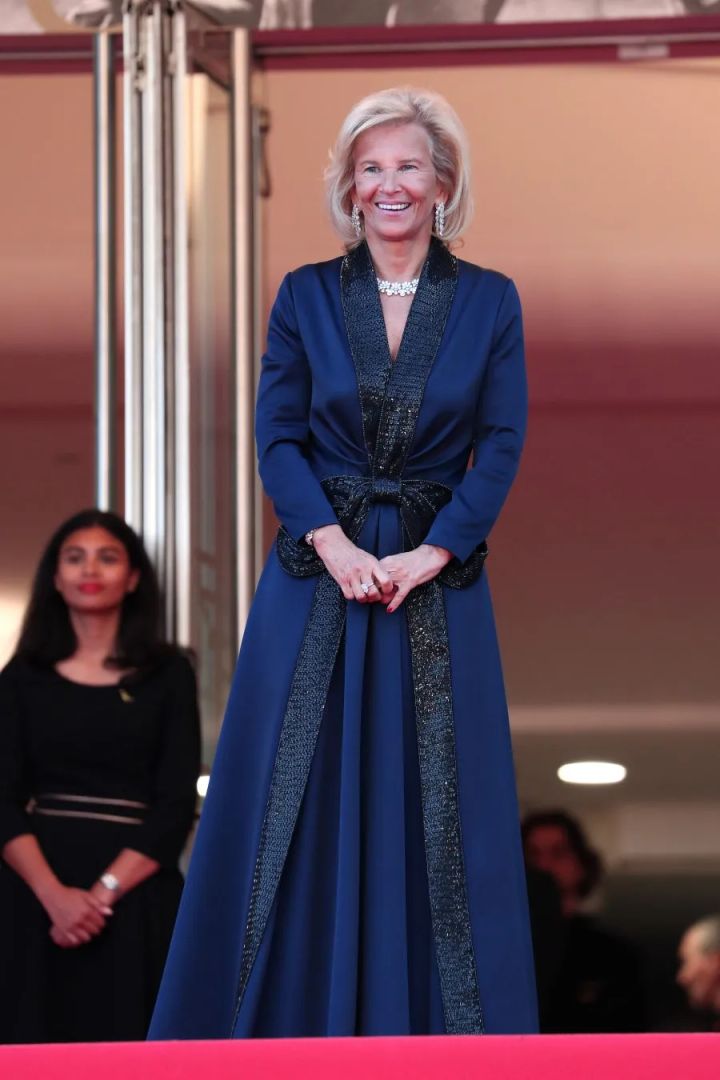 イリス・ノブロックがカンヌ映画祭の会場でブルーのロングコートドレス（黒ラメ刺繍トリム・ウエストリボン）とダイヤモンドネックレスを着用した正面全身ショット