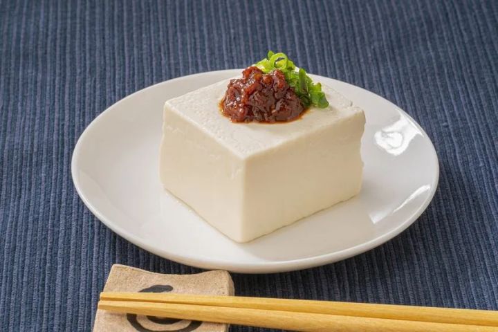白い皿に盛った冷奴の上に旨辛おかず調味料をのせ、小口切り青ねぎを添えた夏向け冷製アレンジ料理の盛り付け写真。手前に箸と箸置きあり。