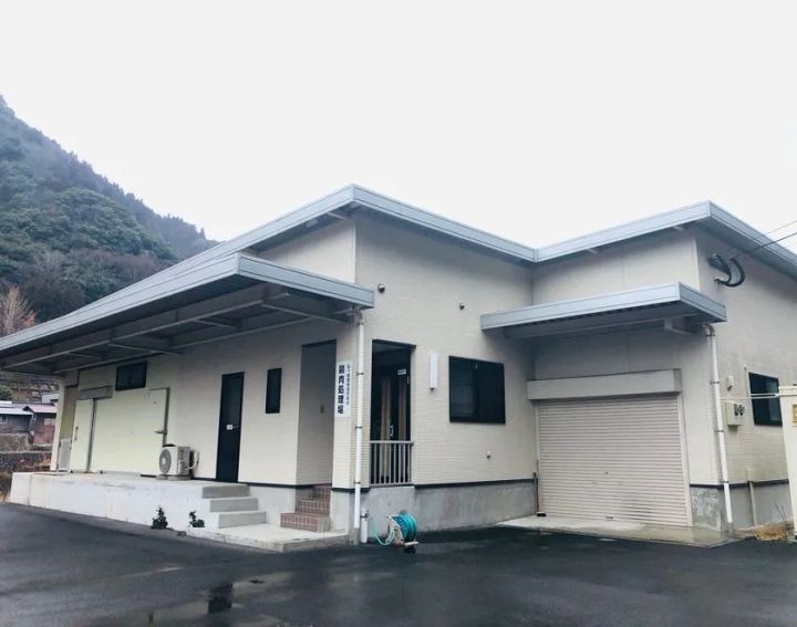 下郷農協から買収した食鳥処理場の外観写真。白壁の平屋建て施設で入口に「鶏肉処理場」の表記があり、シャッター付き搬入口と山を背景にした立地が確認できる