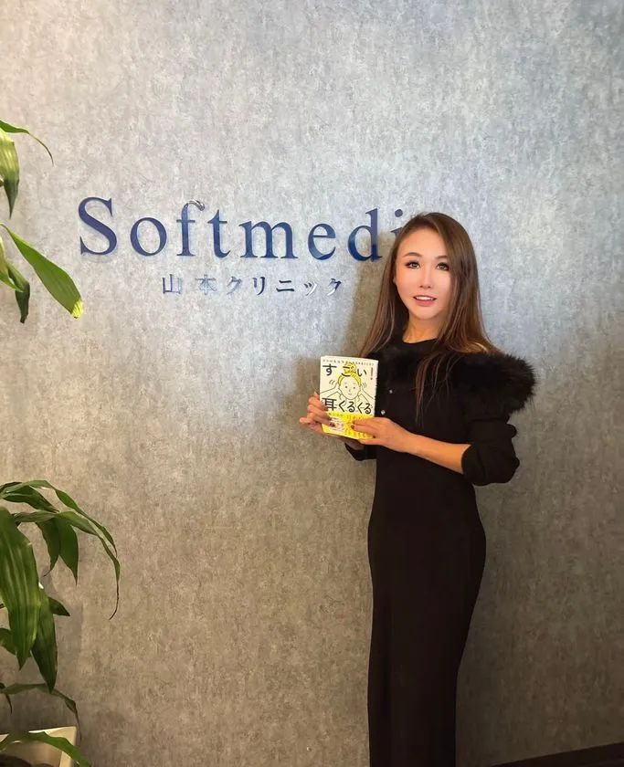 著者・山本江示子が黒ワンピース姿でSoftmedi山本クリニックのロゴ前に立ち、書籍『すごい！耳くるくる』を手に持つポートレート写真