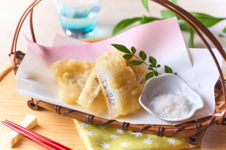穴子天ぷら数切れを竹籠に盛り付け、塩と薬味を添えた料理写真。断面から穴子の身が見えるサクサク感のある揚げ物で、赤い箸と青いガラスコップが夏の演出をしている