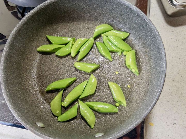 グレーのフライパンで斜め切りのスナップエンドウをサラダ油で炒めている俯瞰写真