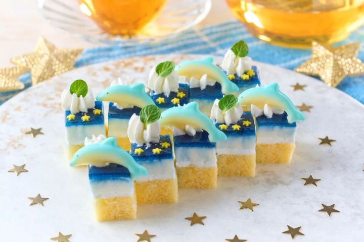 イルカのチョコ飾りとホイップクリーム・ミントを乗せた青×白×黄色のレイヤーゼリーケーキ（一口サイズ）が6個並び、テーブルには金色の星型コンフェッティとティーカップが添えられた夏季限定デザートの商品写真