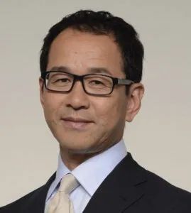 黒縁メガネをかけた40〜50代と思われる男性登壇者の単体プロフィール写真。明るいベージュ背景でスーツ・ネクタイ姿の上半身ショット