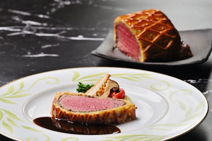 牛フィレ肉のウェリントン包み焼きのスライス断面を金縁白磁皿に盛り付け、後景に包み焼きの全体ブロックを配置した料理写真