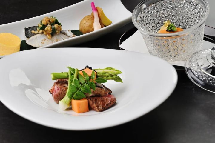 グランキュイジーヌ・コースの複数料理を俯瞰した盛り付け写真。手前の白皿に牛肉とアスパラ・人参・三つ葉を添えたメイン、奥に魚料理と野菜の前菜プレート、右にクリスタルガラス器の冷製スープが並ぶ