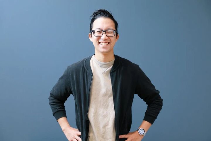 黒いジャージとベージュTシャツを着た眼鏡の男性（登壇者・柴本康平氏）が両手を腰に当てて正面を向いたポートレート写真。グレーブルーの無地背景。