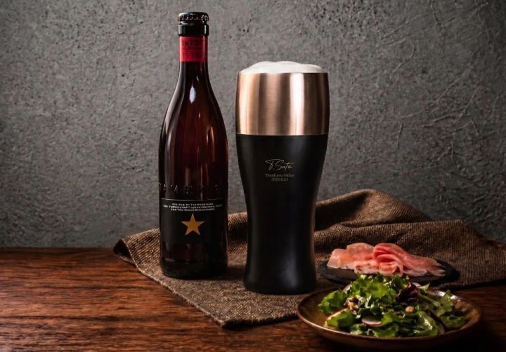 イネディットビール瓶と名入れ刻印入りブラック×ゴールドの真空二重タンブラー（泡あり）の並びショット。手前にサラダと生ハム皿、麻布を配置したテーブルスタイリング