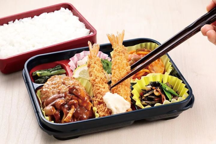玉子屋の弁当容器にエビフライ・ハンバーグ・煮物などの副菜が詰められた仕切り式弁当を箸でつまむアップショット。白飯容器が左奥に分かれて配置されている