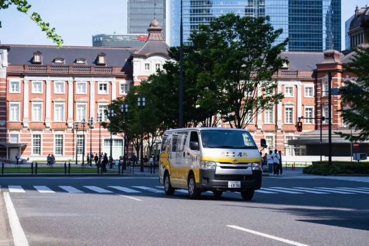 玉子屋のロゴと黄色帯デザインが入ったトヨタ・ハイエースの配達バンが東京駅丸の内口前の交差点を走行している配達車両ショット