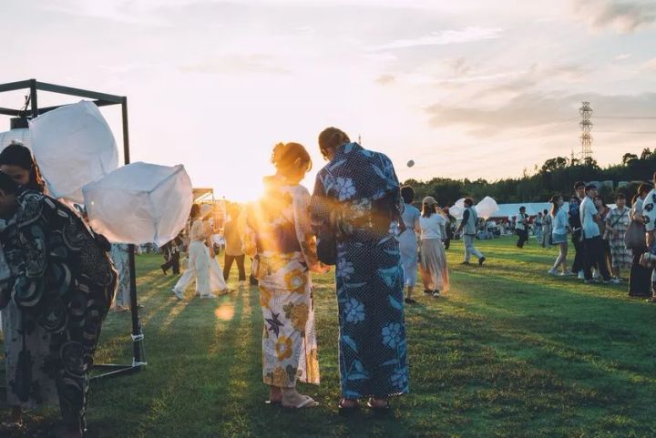 夕暮れ時の広い芝生会場で浴衣姿の参加者カップルがスカイランタンの準備をする後ろ姿。会場全体に多数の来場者が広がり開催規模と参加体験のイメージが伝わる