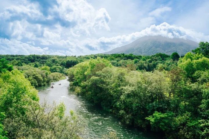 ニセコエリアの緑豊かな夏の川とカヌーを楽しむ人々を俯瞰で捉えた風景写真。背景に雲をまとった山頂が見える