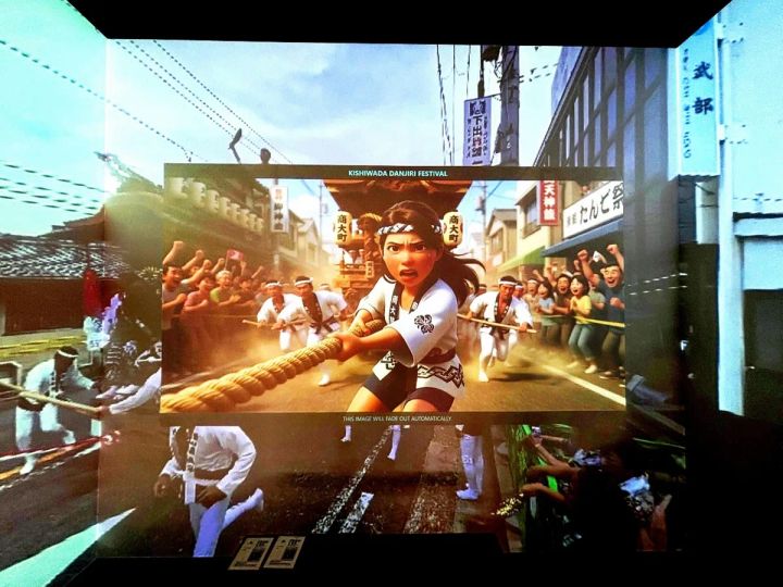 イマーシブ映像体験空間の壁面スクリーンに映し出された岸和田だんじり祭のAI生成アニメ映像で、法被姿の女性キャラクターがだんじりの綱を引く場面と周囲の観衆が描かれている