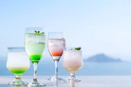 海を背景にテラス席に並ぶ4種のメロン系カクテル・ドリンク。緑・ピンク・白のグラスが並び伊良湖の海景色とともに撮影されたドリンクビジュアル