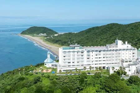 伊良湖オーシャンリゾートの白い高層ホテル建物と礼拝堂を緑の丘と海岸線・青い海を背景に俯瞰で捉えた外観全景写真