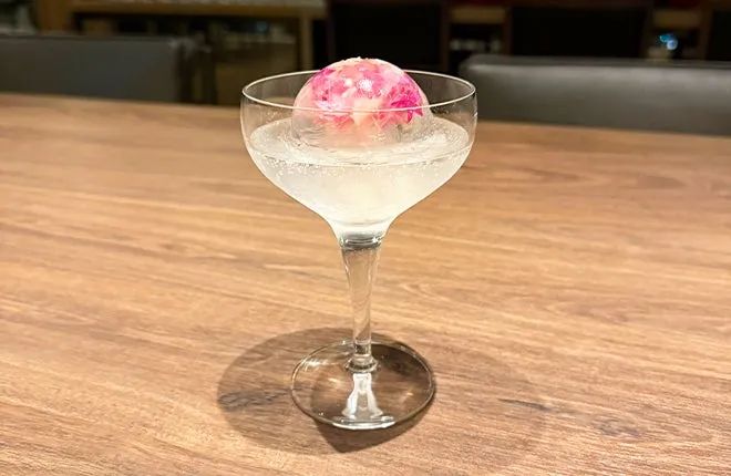 クープグラスに透明な炭酸ベースのドリンクと球形の桜花びら模様ピンクアイスボールを浮かべた20周年限定カクテルの単品クローズアップショット