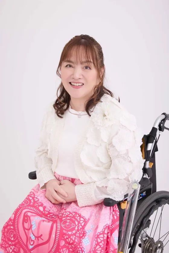 白いレース装飾のブラウスとピンクスカートを着た女性歌手が車椅子に座った上半身アーティスト写真