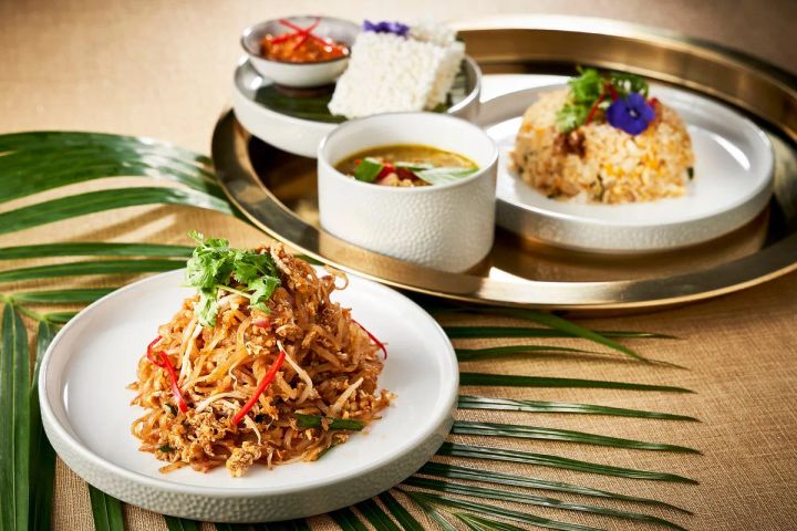 パッタイ（炒め米麺）、グリーンカレー、炒飯、ライスなど複数のタイ料理がシルバートレーと白皿に盛り付けられた料理集合ショット