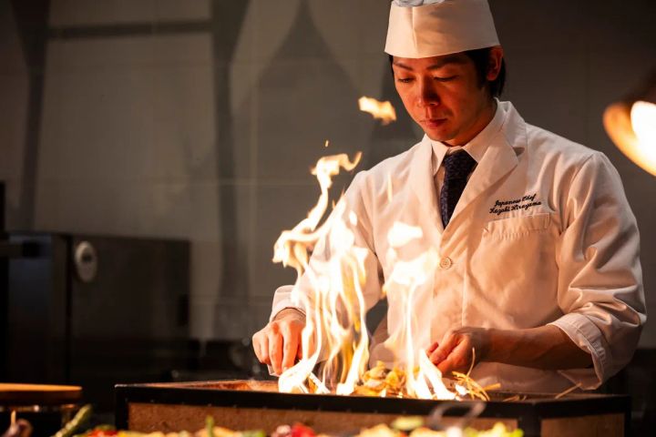 Japanese Chef Kazuki Hirayama と刺繍されたコック服の料理人が鉄板上で大きな炎を上げながら調理するライブクッキングショット