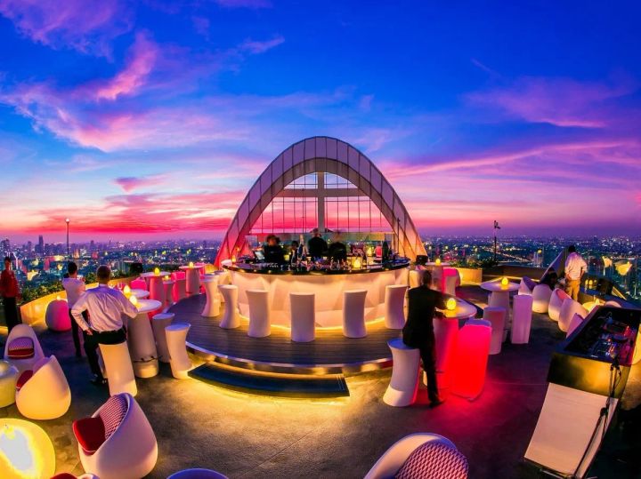 センタラグランドホテル系列のルーフトップバー全景。夕暮れ時の赤紫の空を背景に、アーチ型ガラス構造のバーカウンターと白いイルミネーションチェアが並び、遠方に都市の夜景が広がる。