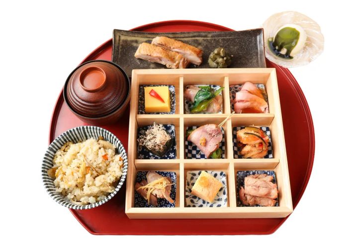 赤い丸盆に乗った九升格子の木箱弁当。鶏・鴨の焼き物・串焼き・煮物・揚げ物など9種の小鉢と、土鍋蓋付きの吸い物、炊き込みご飯、デザートが一膳に揃う夏膳セット