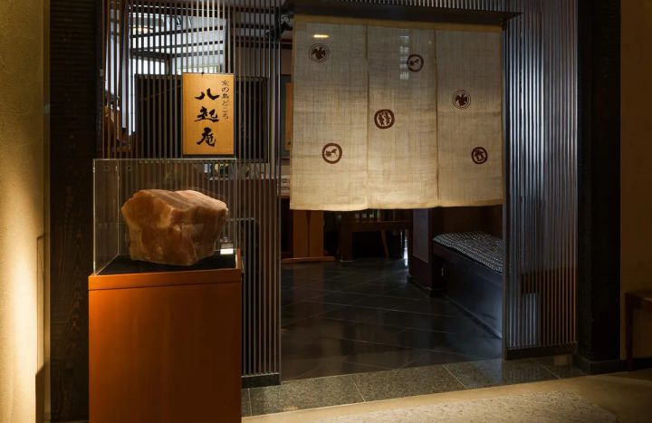 京の鳥どころ八起庵の店舗入口。木製看板と家紋入り白のれん、石塊を展示したガラスケース、格子スクリーンと間接照明で構成された店構えの全景写真