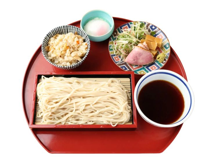 赤い丸盆に乗った鴨つけそば膳。ざる盛りの白い手打ち蕎麦、鴨ロース・メンマ・薬味を盛った鉢、濃いめのつけ汁、炊き込みご飯、塩の小皿で構成された夏限定そばセット