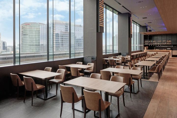 REI Caféとなる9階ラウンジの室内写真。木製テーブルとベージュ革張りチェアが並び、天井まで届く大窓越しにみなとみらいのビル群が一望できる開放的な空間。右奥にバーカウンターが見える。