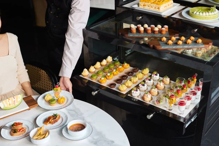 スタッフがワゴンを客席へ引き出しスイーツをサーブする場面。ワゴン上段にケーキ・カヌレ・シュー、中段にフルーツタルト・チョコカップ、下段にムース小瓶が並び、テーブルにはセイボリー皿も置かれる