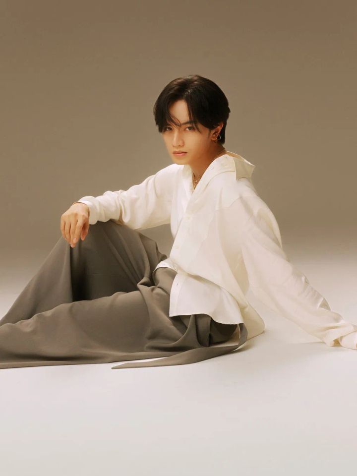 中島健人さんがグレーのワイドパンツと白シャツ姿でスタジオの床に座り、カメラを見つめるポートレート写真。ゴールドのネックレスとピアスを着用。
