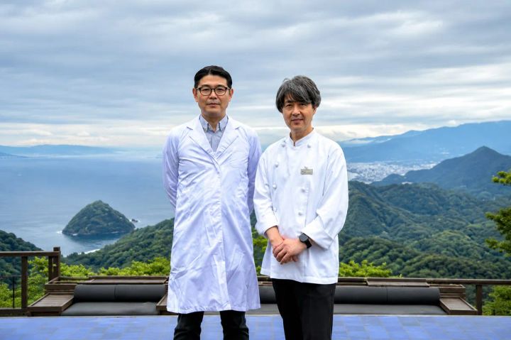 白衣着用の研究者風の男性とシェフコート姿の男性2名が、伊豆の山並みと海を望む高台テラスで並んで立つ連携先担当者の対面写真