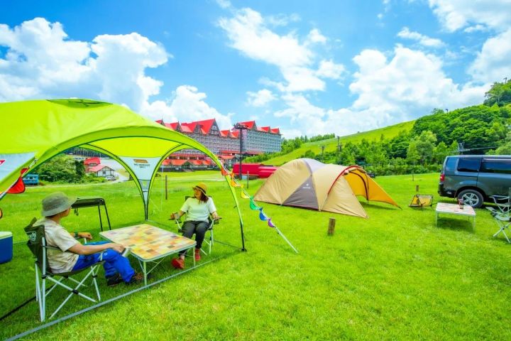 白馬コルチナスキー場の夏ゲレンデにテントとタープを設営し、ホテル棟を背景に親子がキャンプチェアでくつろぐグランピング風景