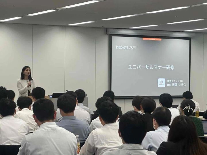 株式会社ノジマのユニバーサルマナー研修の開講場面。ミライロ講師・薄葉ゆきえ氏がスクリーン前でマイクを持ち登壇し、白シャツ姿の多数の社員が着席して受講している様子。