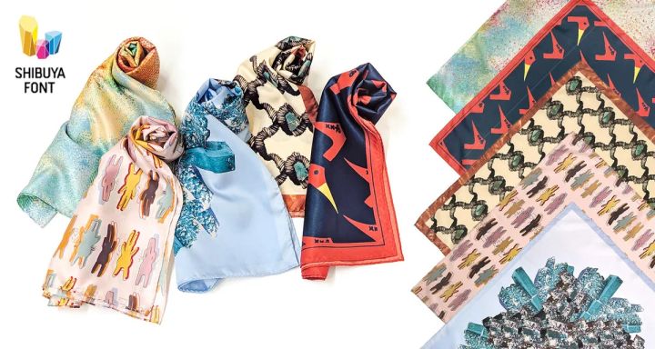 シブヤフォント×サーヴォのコラボスカーフ全5種を一覧で撮影した商品写真。ロールして立てた3本と平置き重ねた5枚を並べ、人物柄・鳥柄・抽象柄など各デザインの色味と柄の違いを比較できる構成。