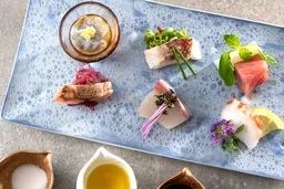 淡青色の石皿に盛られた鮮魚や野菜の前菜盛り合わせ。複数の小皿（塩・オイル・ソース類）が添えられた日本料理コースの一皿で、花びら・緑葉・薄切り刺身などが丁寧に配置されている。