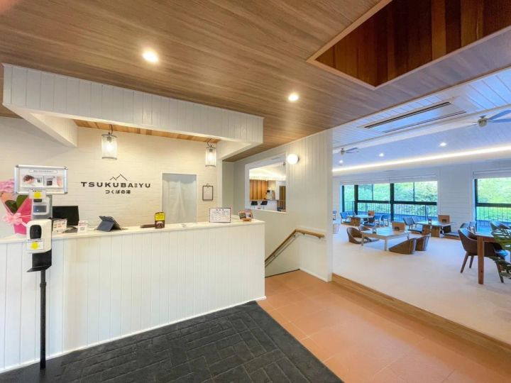つくばの湯フロント受付カウンターと奥に広がる窓際カフェスペース。木目天井・白壁の内装で、窓外に緑が見える明るい開放的な空間構成