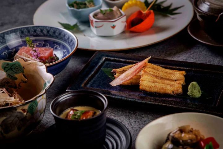 日本料理コースの卓上全景。中央の黒長皿に鰻白焼きとわさびを配し、茶碗蒸し・刺身・前菜小鉢・焼き物など複数料理が一同に並ぶ会席スタイルの盛り付け写真